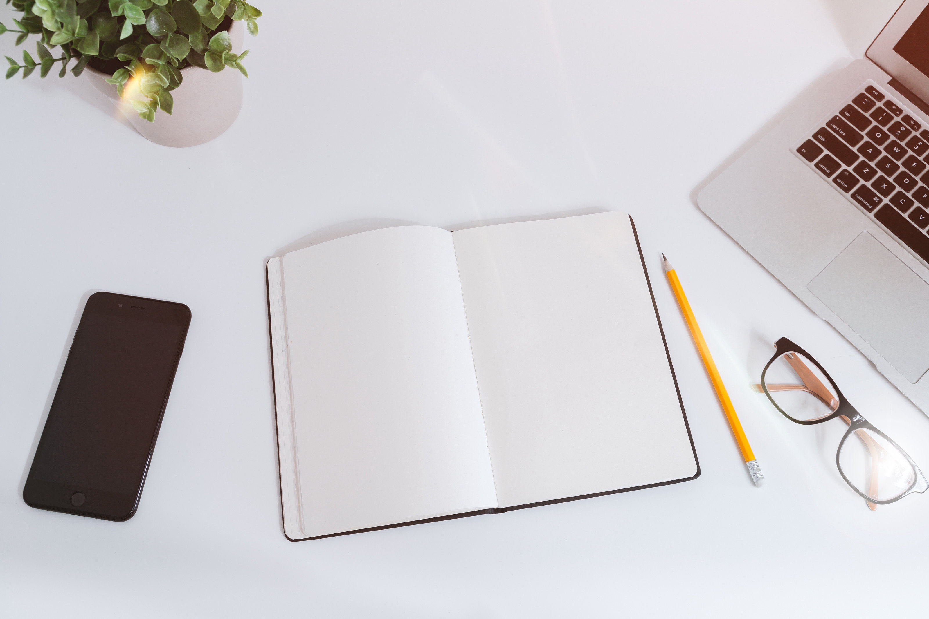 Smart phone, plant, computer, glasses, and open paper journal on a white table